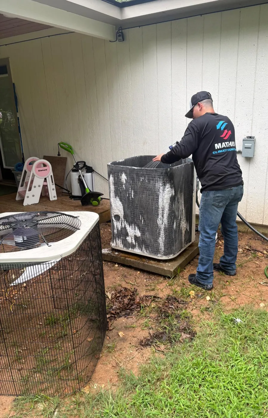 Technician in a Mathis Climate Control shirt cleaning an outdoor condenser in a backyard